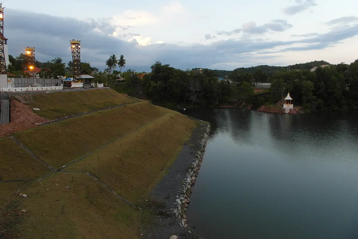 A grassy bank and lit lantern towers beside the lake at Siam Niramit Phuket, with a small gold shrine on the far shore