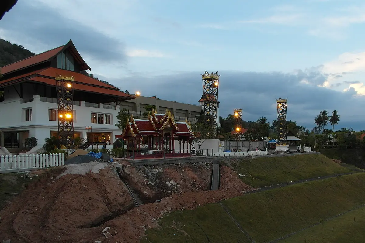 A red and gold Thai pavilion in the Siam Niramit Phuket grounds at dusk, with a large white building and lit lantern towers behind